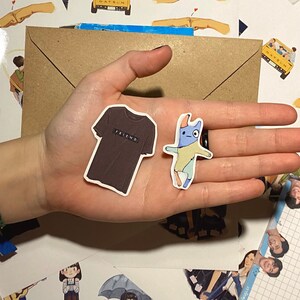 Op de afbeelding: Een hand die twee stickers vasthoudt. E&eacute;n sticker is een bruin T-shirt met het woord "FRIEND" erop gedrukt. De andere sticker is een cartoon van een blauw en geel wezen met een rode neus.