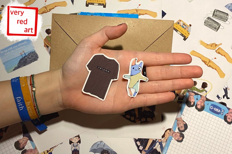 Op de afbeelding: Een hand die twee stickers vasthoudt. E&eacute;n sticker is een bruin T-shirt met het woord "FRIEND" erop gedrukt. De andere sticker is een cartoon van een blauw en geel wezen met een rode neus.