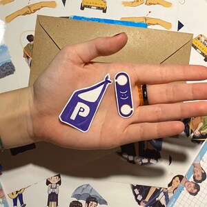 Op de afbeelding: Twee stickers, een paarse met een witte "P" en een witte tasvorm, de andere blauw met een witte smiley, op een hand met een blauwe armband waarop "faith happy" staat.