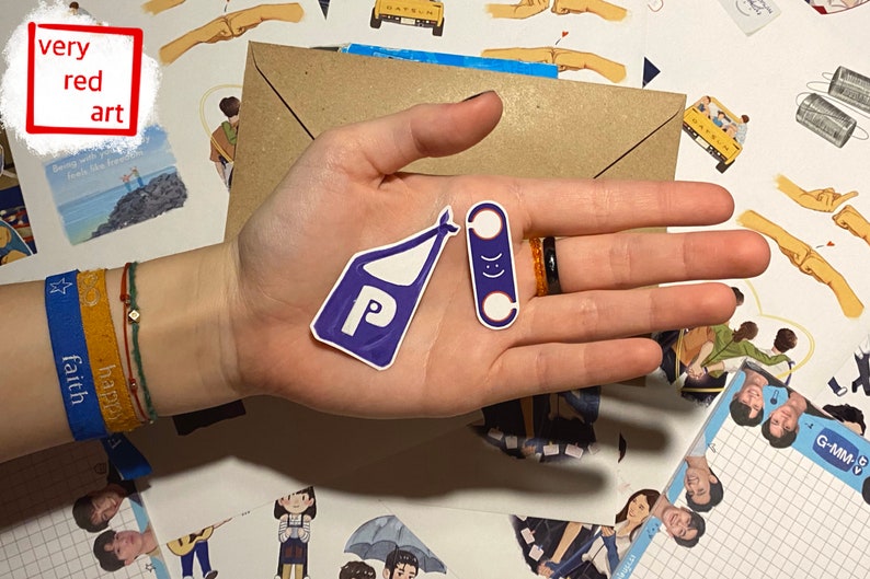 Op de afbeelding: Twee stickers, een paarse met een witte "P" en een witte tasvorm, de andere blauw met een witte smiley, op een hand met een blauwe armband waarop "faith happy" staat.