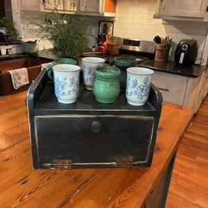 May include: A black wooden bread box with a drop-down front door and a single knob, topped with two white mugs featuring blue floral patterns, one green ceramic jar, and one dark green mug, all arranged on a wooden kitchen countertop.