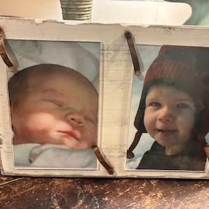 May include: A distressed white wooden double picture frame with two photos. One photo shows a newborn baby, and the other shows a toddler wearing a red hat. The frame has copper-colored accents.