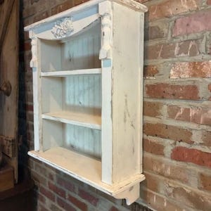 May include: A distressed white wall-mounted shelf unit with three shelves. The top features a decorative floral carving and a light blue accent. The unit is mounted on a red brick wall, showcasing a vintage aesthetic.
