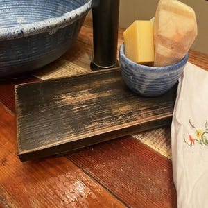May include: A rectangular black wooden riser with a distressed finish, holding a small blue ceramic bowl containing a rectangular yellow soap bar and a larger, irregular, light-colored textured soap block with visible orange-toned mineral veins.