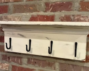 May include: A wall-mounted shelf featuring a distressed white finish with visible wood grain and wear. The unit includes a flat top ledge and four evenly spaced black metal hooks attached to the front panel for hanging various items securely.