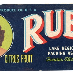 May include: A vintage citrus fruit label featuring a woman's portrait in a circle, the word "RUBY" in large, stylised letters, and the text "PRODUCE OF U.S.A. CITRUS FRUIT LAKE REGION PACKING ASSN. Tavares, Florida BRAND".