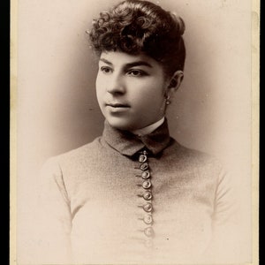 Puede incluir: Una fotografía en blanco y negro de una mujer con el pelo rizado que lleva una blusa con cuello y una fila de botones en la parte delantera. La foto es del estudio "Huszagh" ubicado en 163 S. Halsted St., Chicago.