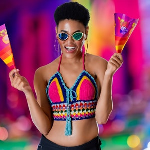 May include: A woman wearing a colorful crocheted top and sunglasses holds two small flags with colorful designs. The background is blurred and features a variety of colors.