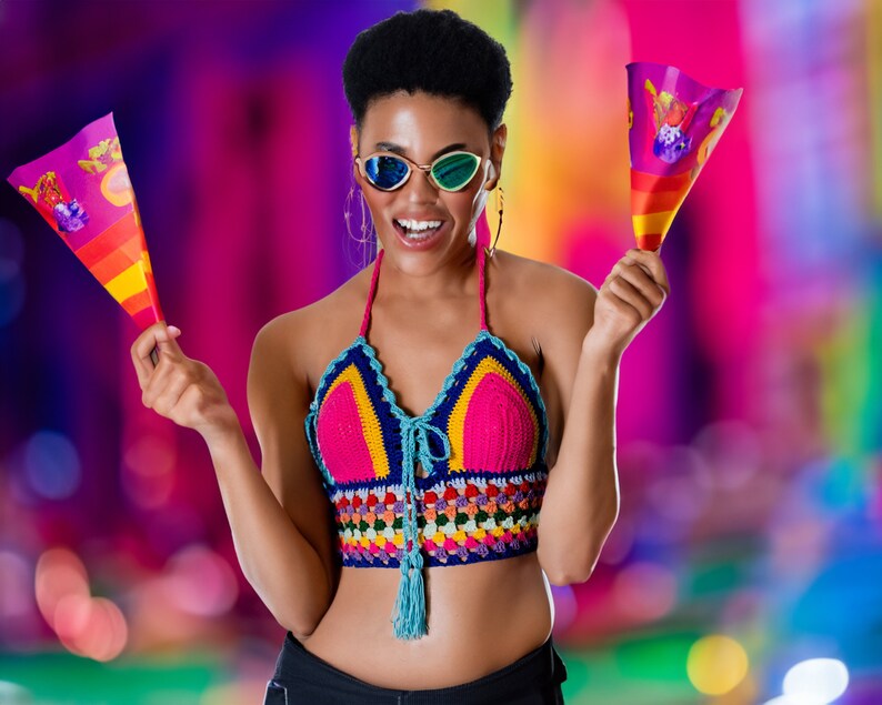 May include: A woman wearing a colorful crocheted top and sunglasses holds two small flags with colorful designs. The background is blurred and features a variety of colors.