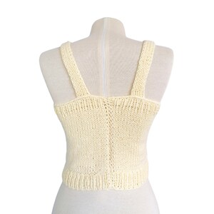 May include: A cream-colored knitted tank top with a scoop neck and thin straps. The top is made of a soft, lightweight yarn and has a relaxed fit.