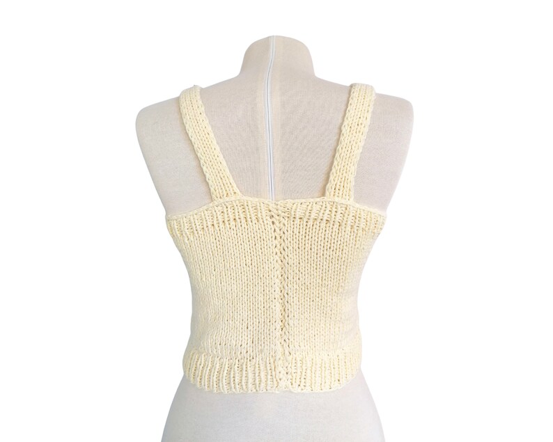 May include: A cream-colored knitted tank top with a scoop neck and thin straps. The top is made of a soft, lightweight yarn and has a relaxed fit.
