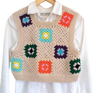 Op de afbeelding: Een beige gebreide vest met een kleurrijk granny square-patroon. Het vest heeft een ronde hals en is mouwloos. De vierkanten zijn in verschillende kleuren, waaronder groen, blauw, roze, oranje en geel.