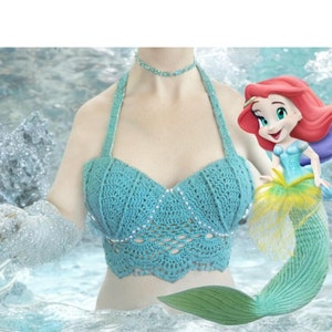 May include: A teal crochet top with a mermaid tail and a cartoon mermaid illustration. The top has a scalloped edge and is decorated with small, sparkly beads.