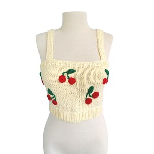May include: A cream-colored knitted crop top with red cherries embroidered on it. The top has thin straps and a square neckline.