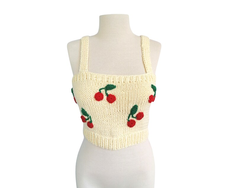 May include: A cream-colored knitted crop top with red cherries embroidered on it. The top has thin straps and a square neckline.