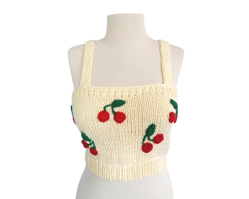 May include: A cream-colored knitted crop top with cherry embroidery. The cherries are red with green stems and leaves.