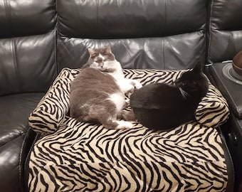 May include: Two cats, one gray and white and one black, are relaxing on a zebra-print cat bed. The bed is on a black leather couch.