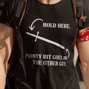 May include: A black t-shirt with a white graphic of a sword and the text "Hold Here. Pointy bit goes in the other guy."