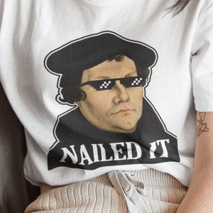 May include: White t-shirt featuring a black and white image of a man wearing a black hat and sunglasses with the text "NAILED IT" underneath.