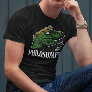 May include: A black t-shirt featuring a green dinosaur wearing a gold laurel wreath on its head. The text "PHILOSORAPTOR" is printed in white below the dinosaur.