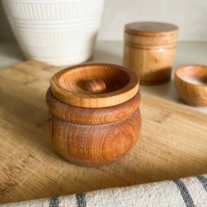 May include: A set of three wooden containers with lids. The containers are made of light brown wood and have a natural finish. The lids are round and have a small knob on top. The containers are sitting on a wooden cutting board.