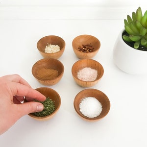 May include: Six small wooden bowls filled with various spices and seasonings, including salt, pepper, herbs, and dried chili flakes. The bowls are arranged in two rows of three on a white surface.