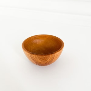 May include: A small, wooden bowl with a natural wood finish. The bowl is empty and has a smooth, rounded shape.