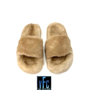 May include: A pair of beige faux fur sliders. The sliders have a wide strap across the top of the foot and a simple, open-toe design.