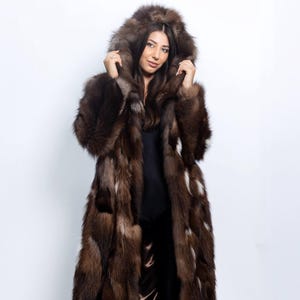 May include: A full-length fur coat in brown tones with a hood. The coat features white highlights and is worn over black clothing. The text "VFC" is visible. This coat is a luxurious fashion item, suitable for colder weather.