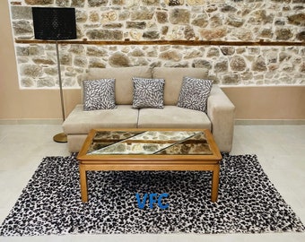 Puede incluir: Un sofá beige con tres almohadas se encuentra frente a una pared de piedra. Una mesa de café de madera con tapa de vidrio se encuentra sobre una alfombra de estampado animal negro y blanco. La alfombra está frente al sofá.
