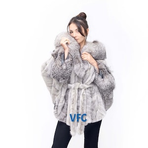 May include: A silver fox fur coat with a large collar and a belt. The coat is worn by a person with black trousers and silver heels. The text "VFC" is visible on the coat.