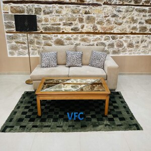 Pode incluir: Uma sala de estar com um sof&aacute; bege, tr&ecirc;s almofadas com padr&atilde;o floral, uma mesa de centro de madeira com tampo de vidro e um tapete verde com padr&atilde;o xadrez. A parede atr&aacute;s do sof&aacute; &eacute; feita de pedra.