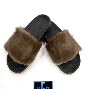 May include: A pair of brown fur slides with black soles. The slides are made of real fur and have a comfortable fit.
