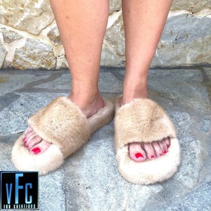 May include: A pair of beige fur slippers with a fluffy texture. The slippers have a simple design with a wide strap across the top of the foot.