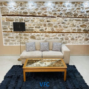 May include: A beige sofa with three throw pillows sits in front of a stone wall. A black lamp with a patterned shade stands next to the sofa. A dark blue shag rug covers the floor in front of the sofa. A wooden coffee table with a glass top sits on the rug. The coffee table has a mirrored top with a geometric pattern. The text "VFC" is printed on the rug.
