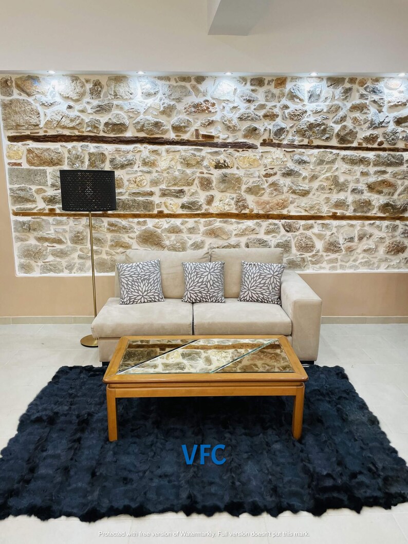 May include: A beige sofa with three throw pillows sits in front of a stone wall. A black lamp with a patterned shade stands next to the sofa. A dark blue shag rug covers the floor in front of the sofa. A wooden coffee table with a glass top sits on the rug. The coffee table has a mirrored top with a geometric pattern. The text "VFC" is printed on the rug.