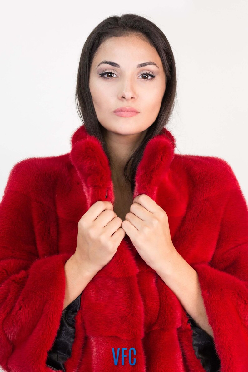 Peut inclure: Une femme portant un manteau en fourrure rouge avec un logo VFC en bas. Elle tient le col du manteau ferm&eacute;.