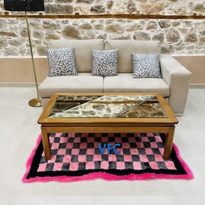 Pode incluir: Uma sala de estar com um sofá bege, três almofadas decorativas e uma mesa de centro de madeira com tampo de pedra e vidro. Um tapete xadrez rosa e preto está em frente. Uma luminária de chão preta é visível.