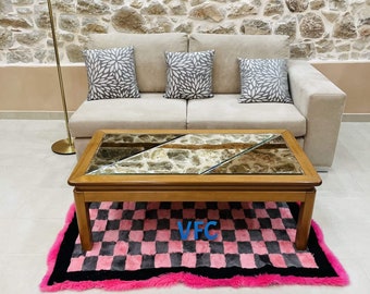 Puede incluir: Un salón con un sofá beige, tres cojines decorativos y una mesa de centro de madera con tapa de piedra y cristal. Una alfombra a cuadros rosa y negra está delante. Se ve una lámpara de pie negra.