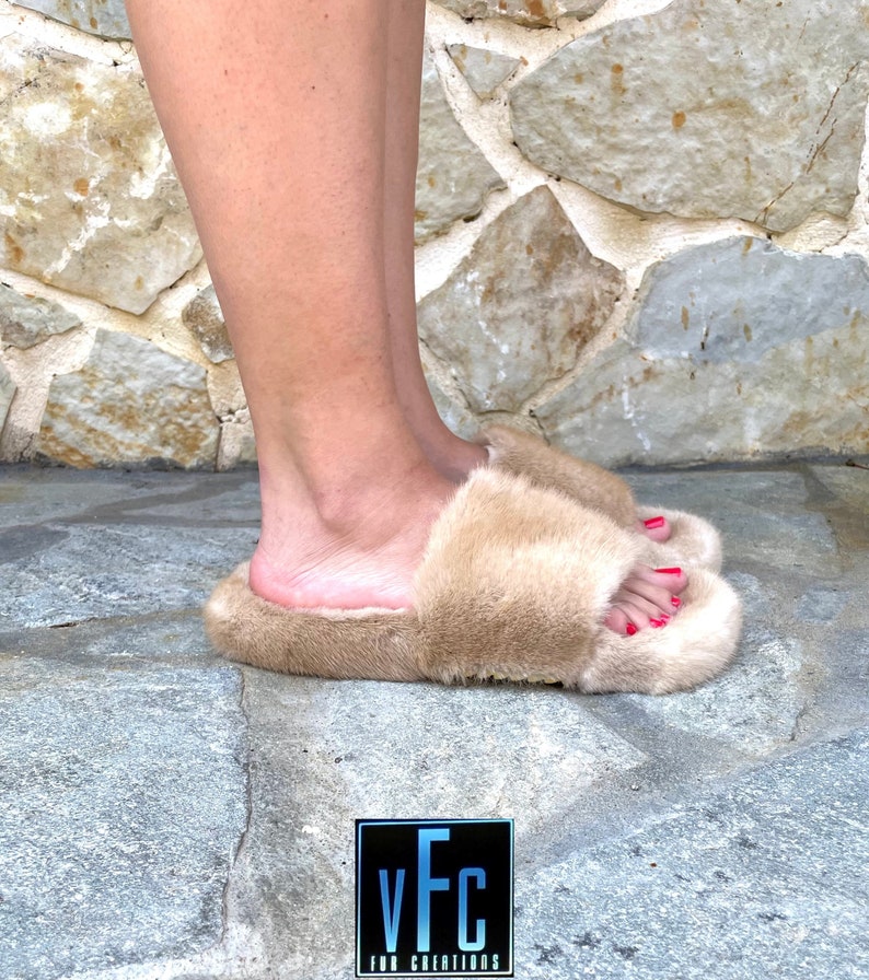 May include: A pair of beige fur slides with a square toe. The slides are made of soft, fluffy fur and have a simple, elegant design.
