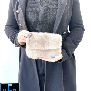 May include: A small, light brown fur clutch bag. The bag is being held by a person wearing a black turtleneck jumper and a black coat.