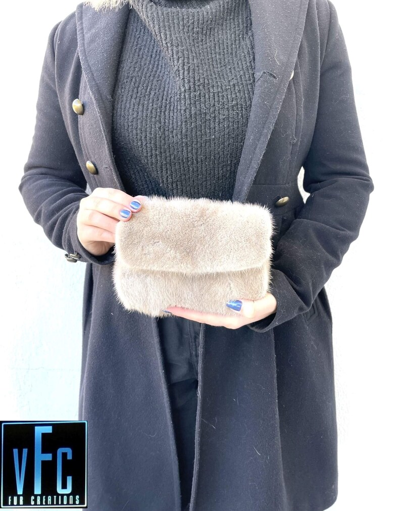 May include: A small, light brown fur clutch bag. The bag is being held by a person wearing a black turtleneck jumper and a black coat.