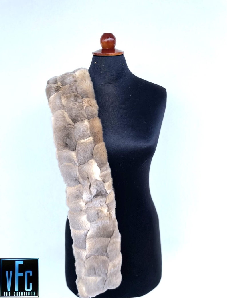 May include: A long, light brown fur scarf with a braided design. The scarf is draped over a black mannequin.
