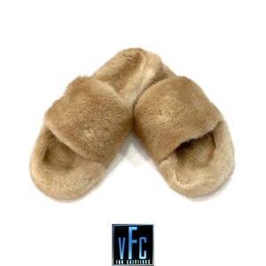 May include: A pair of tan fur slides. The slides are made of soft, fluffy fur and have a simple, slip-on design.