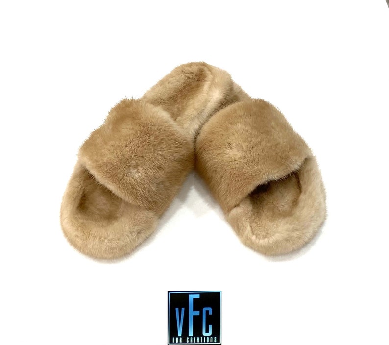 May include: A pair of tan fur slides. The slides are made of soft, fluffy fur and have a simple, slip-on design.