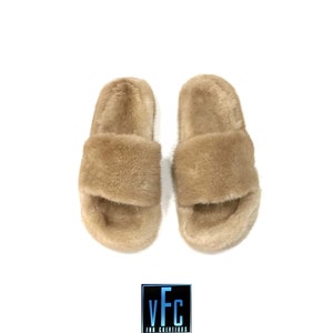 May include: A pair of light brown fur slides. The slides have a wide strap across the top of the foot and a fluffy, soft texture.