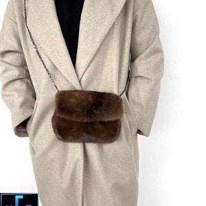 May include: A brown fur crossbody bag with a silver chain strap. The bag is worn by a person wearing a beige coat.