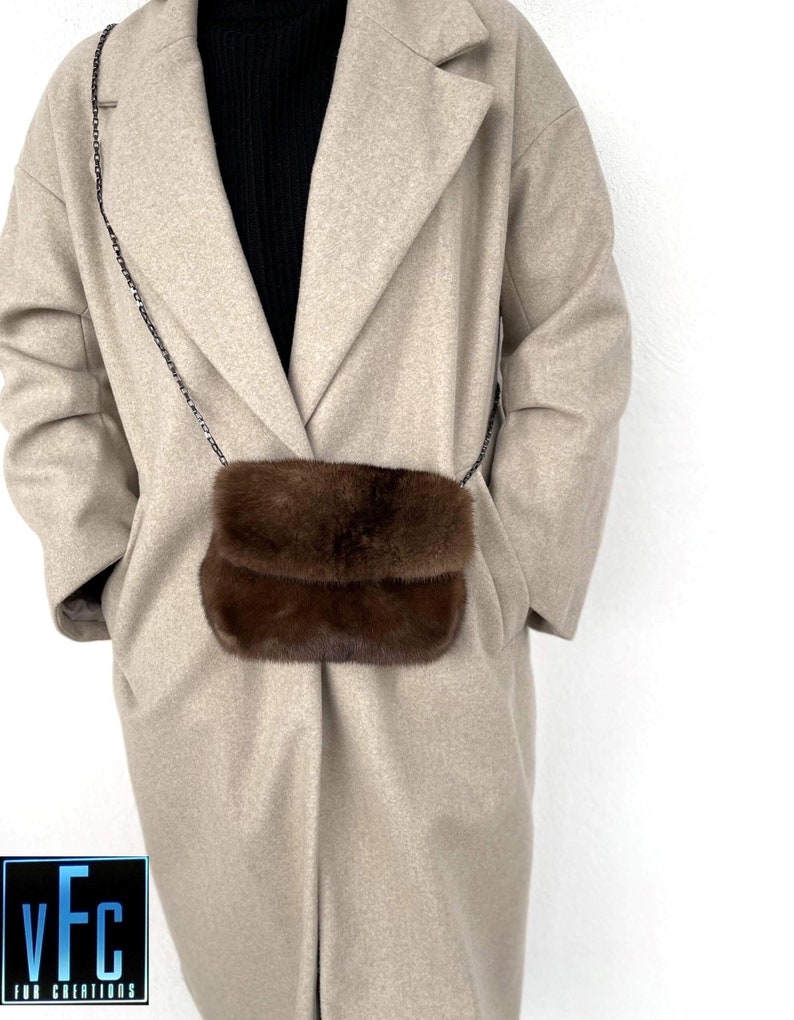 May include: A brown fur crossbody bag with a silver chain strap. The bag is worn by a person wearing a beige coat.