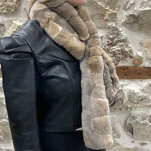 May include: A brown faux fur scarf with a long, rectangular shape. The scarf is made of soft, fluffy fur and has a subtle pattern. It is worn by a person wearing a black leather jacket.