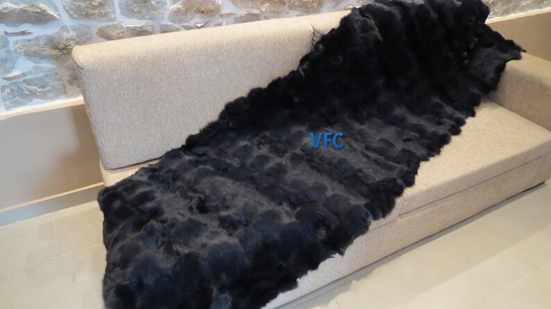 May include: A black rabbit fur throw blanket is draped over a light brown couch. The blanket is made of many strips of fur sewn together. The text "VFC" is visible on the blanket.
